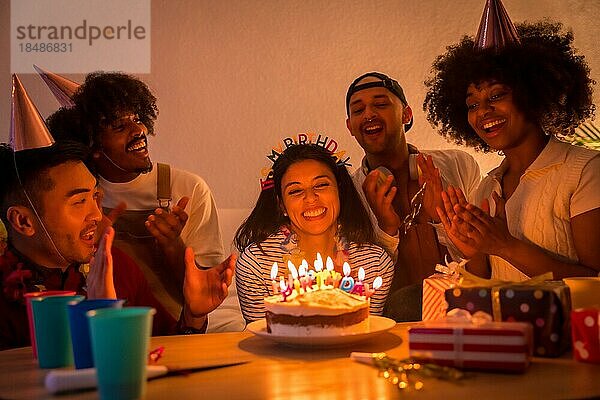 Multiethnische Gruppe von Freunden bei einer Geburtstagsfeier auf dem Sofa zu Hause mit Kuchen und Geschenken  bei ausgeschaltetem Licht  die Happy Birthday singen