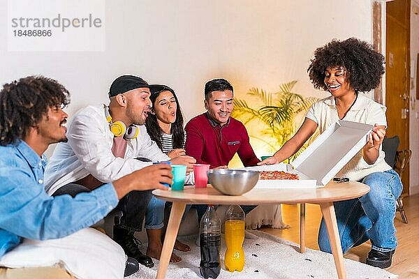 Eine Gruppe multiethnischer Freunde sitzt auf einem Sofa  isst Pizza und trinkt alkoholfreie Getränke bei einer Party zu Hause  ein junger Mann kommt mit einem Pizzakarton von der Straße