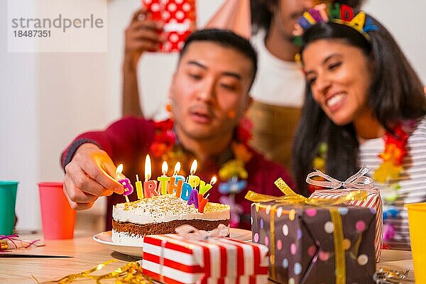 Multiethnische Gruppe von Freunden bei einer Geburtstagsfeier auf dem Sofa zu Hause mit einem Kuchen und Geschenken  Anzünden der Kerzen