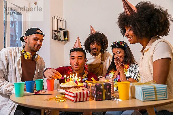 Multiethnische Gruppe von Freunden bei einer Geburtstagsfeier auf dem Sofa zu Hause mit einem Kuchen und Geschenken  beim Anzünden der Kerzen