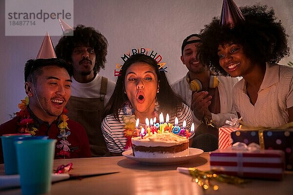 Multiethnische Gruppe von Freunden bei einer Geburtstagsfeier auf dem heimischen Sofa mit Kuchen und Geschenken  Ausblasen der Kerzen bei ausgeschaltetem Licht