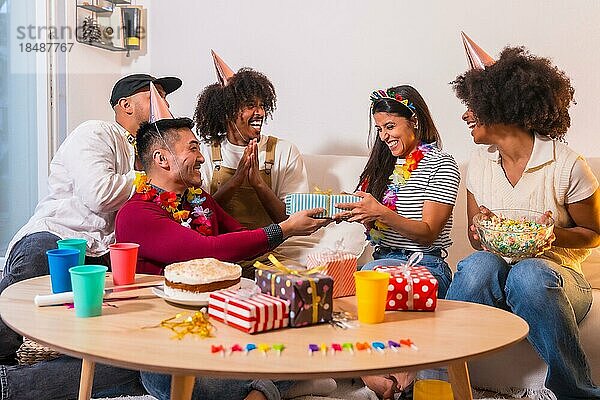 Gruppe von Freunden bei einer Geburtstagsfeier auf dem Sofa zu Hause  die lächelnd die Geschenke überreichen und das Geburtstagskind überraschen