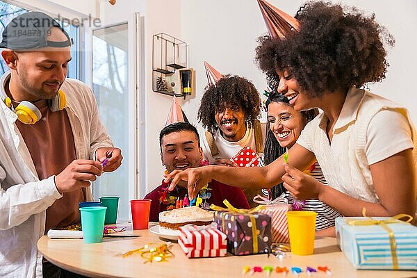 Multiethnische Gruppe von Freunden bei einer Geburtstagsfeier auf dem Sofa zu Hause mit einem Kuchen und Geschenken  Anbringen der Kerzen