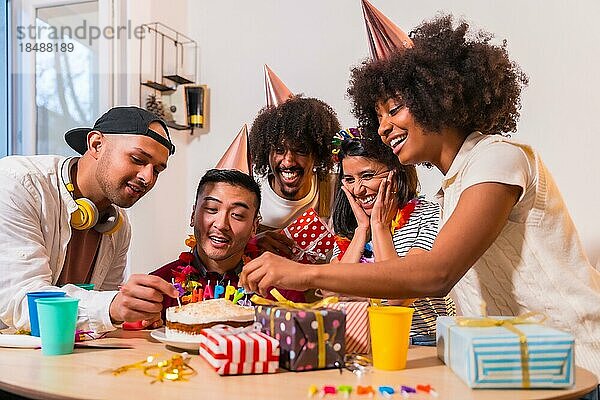 Multiethnische Gruppe von Freunden bei einer Geburtstagsfeier auf dem Sofa zu Hause mit einem Kuchen und Geschenken  lächelnd Platzierung der Kerzen