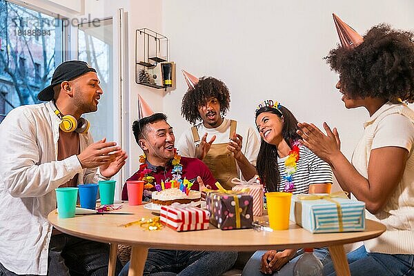 Multiethnische Gruppe von Freunden bei einer Geburtstagsfeier auf dem Sofa zu Hause mit einem Kuchen und Geschenken  die ihm ein Geburtstagsständchen singen