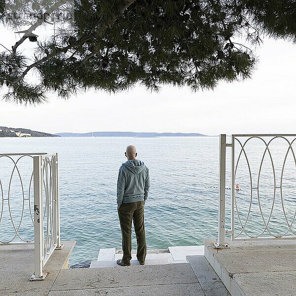 Mann steht am Meer unter einer Pinie und schaut auf das Wasser  Seget Vranjica  Kroatien  Europa
