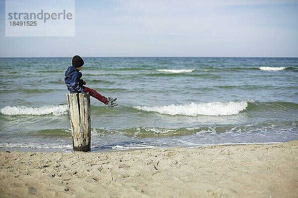 Symbolfoto zum Thema Einsamkeit in der Kindheit. Ein Kind sitzt allein auf einem Holzpfosten am Meer. Arenshoop  20.04.2022  Arenshoop  Deutschland  Europa