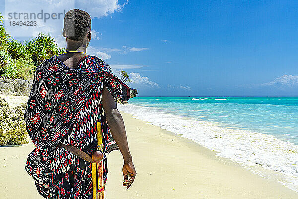 Maasai-Mann mit traditioneller Kleidung und Stock bewundert das kristallklare Meer am Strand  Sansibar  Tansania  Ostafrika  Afrika
