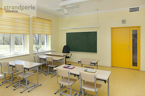 Ein neu gebautes Schulklassenzimmer mit Schreibtisch und Stühlen. Fenster mit gelben Jalousien.