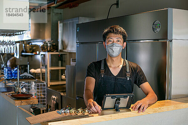 Ein Mann mit Gesichtsmaske steht am Tresen einer Restaurantküche und benutzt ein digitales Tablet  der Besitzer oder Manager.