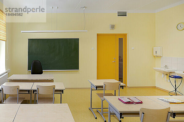Klassenzimmer mit Schreibtisch und Stühlen. Fenster mit gelben Jalousien und grünem Brett.