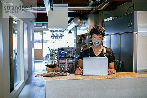 Ein Mann mit Gesichtsmaske in einer Restaurantküche  der einen Laptop benutzt  der Besitzer oder Manager.