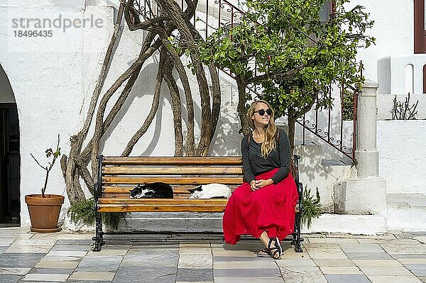 Junger Frau mit rotem Rock sitzt auf einer Bank mit schlafenden Katzen  Kloster Panagia Tourliani  Ano Mera  Mykonos  Kykladen  Griechenland  Europa