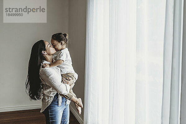 Junge Mutter mit langen Haaren hält Kleinkind im Arm und küsst sich im Studio