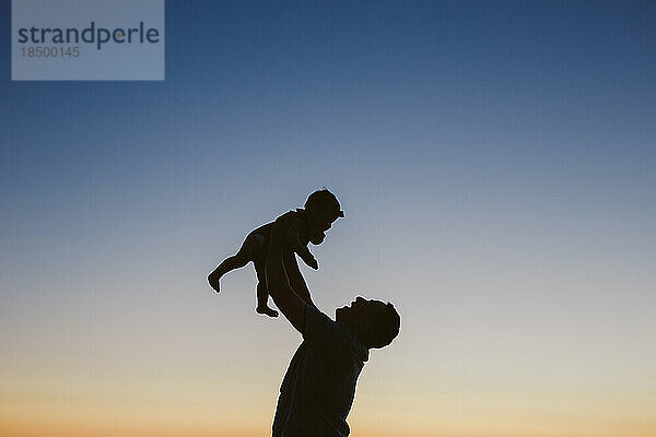 Silhouette eines Vaters  der sein Baby bei Sonnenuntergang in der Luft hält