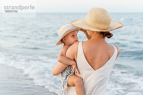 Frau mit Strohhut hält ein kleines Mädchen im Arm am Meer