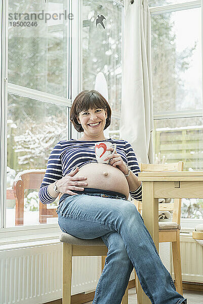 Porträt einer schwangeren Frau  die mit einer Kaffeetasse auf einem Stuhl sitzt