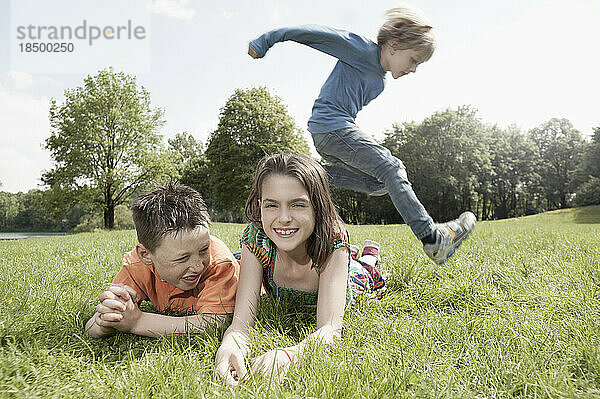 Junge springt über seine auf Gras liegenden Freunde in einem Park  München  Bayern  Deutschland