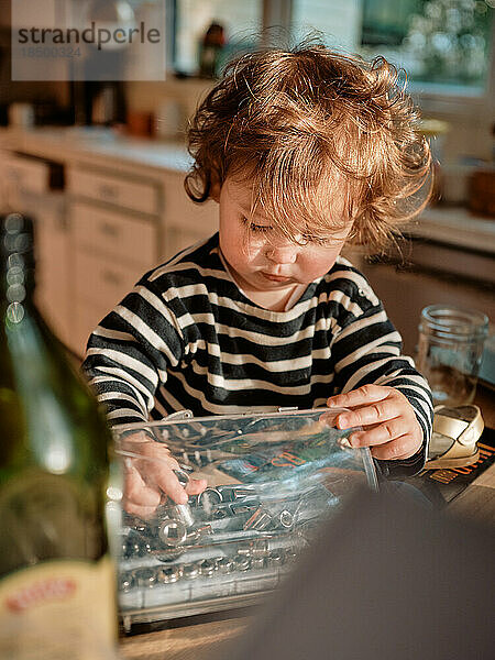 Kleinkind mit lockigen roten Haaren spielt in einer unordentlichen Küche mit Werkzeugen