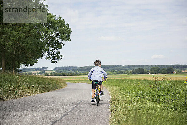 Rückansicht eines Kindes  das Fahrrad auf der Straße auf dem Land fährt  Bayern  Deutschland