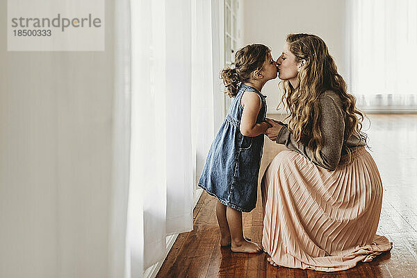 Mother bending down to kiss young daughter in natrual light studio