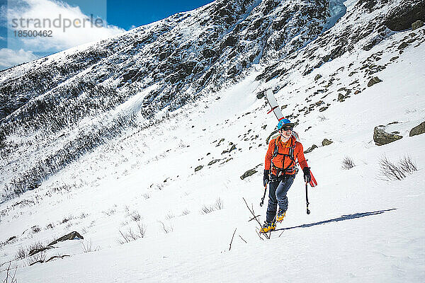 Mann geht mit Eiskletterwerkzeug und Skiern auf dem Rücken bergauf