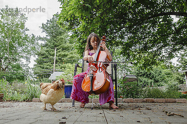 Ein kleines barfüßiges Mädchen spielt Cello für ihr Haustierhuhn im Garten