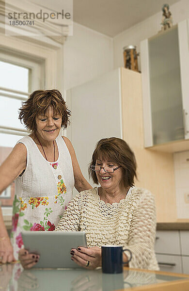 Zwei ältere Mitbewohner nutzen ein digitales Tablet und lächeln in der Küche  München  Bayern  Deutschland