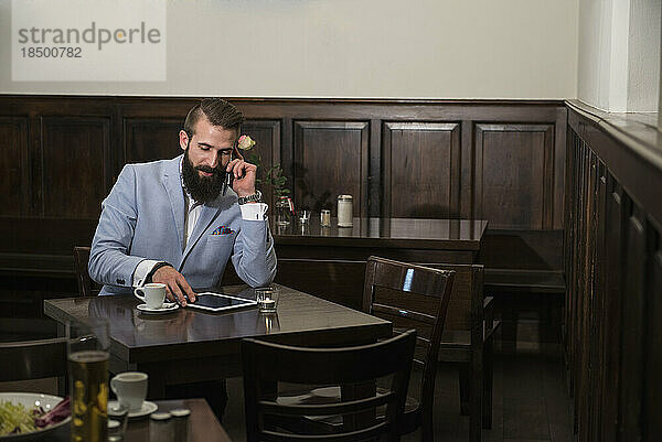 Junger Mann telefoniert  während er im Restaurant ein digitales Tablet nutzt