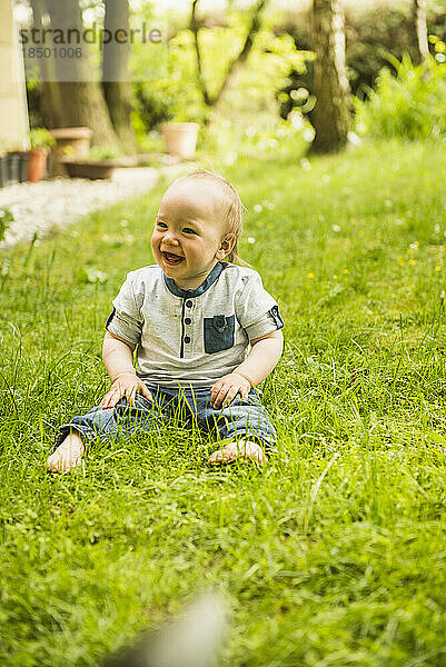 Lachender kleiner Junge sitzt auf der Wiese im Garten
