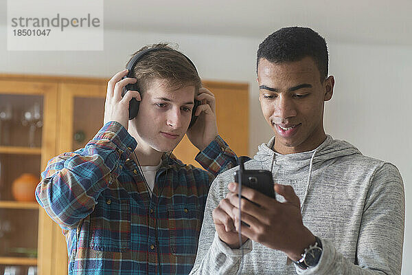 Junger Freund hält Mobiltelefon in der Hand  während ein anderer Freund Kopfhörer trägt und Musik hört