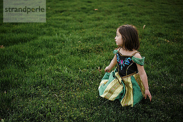 A small girl in a princess costume sits alone in a grassy field