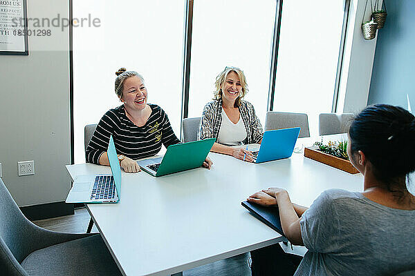 Zwei Frauen lächeln einer dritten Frau zu  während sie an einem Konferenztisch sitzen