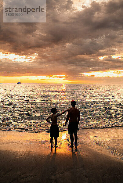 Vater und Sohn stehen an einem Sandstrand und beobachten gemeinsam den Sonnenuntergang.