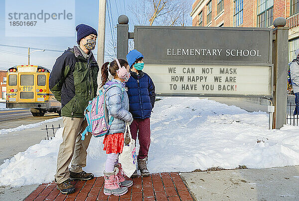 Ein maskierter Vater wartet mit zwei Kindern vor der Grundschule
