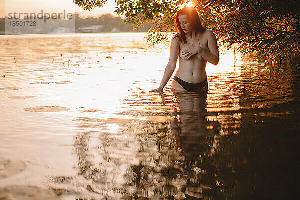 Oben-ohne-Frau bedeckt ihre Brust  während sie bei Sonnenuntergang im See steht