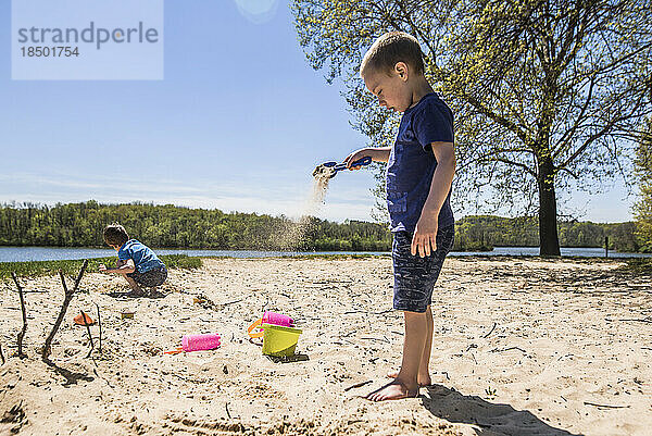 Junge schüttet Sand aus Schaufel in den Wind  während er am Seeufer steht