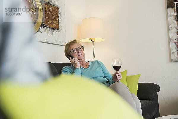 Alte Frau entspannt sich auf dem Sofa bei einem Glas Rotwein und telefoniert