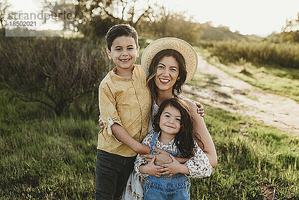 Porträt einer glücklichen jungen Mutter und ihrer Kinder  die auf einer Wiese im Gegenlicht lächeln
