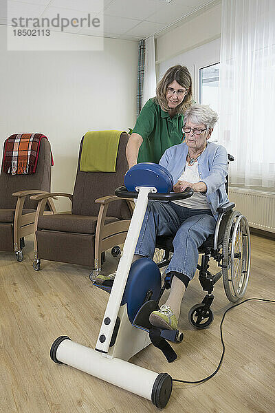 Krankenschwester und ältere Frau im Rollstuhl trainieren auf Heimtrainer im Pflegeheim
