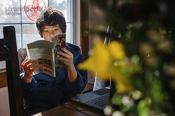 Ein Junge sitzt am Tisch und liest bei Fensterlicht ein Buch