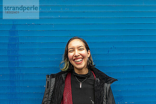 Junge Frau lacht und blickt in die Kamera  steht auf einer blauen Wand.