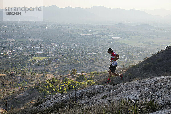 Sportlicher männlicher Trailrunner fliegt felsiges Gelände in den trockenen Bergen von El Arenal  Hidalgo  Mexiko  hinunter  während die Sonne in der Ferne untergeht und die Stadt unten erleuchtet.