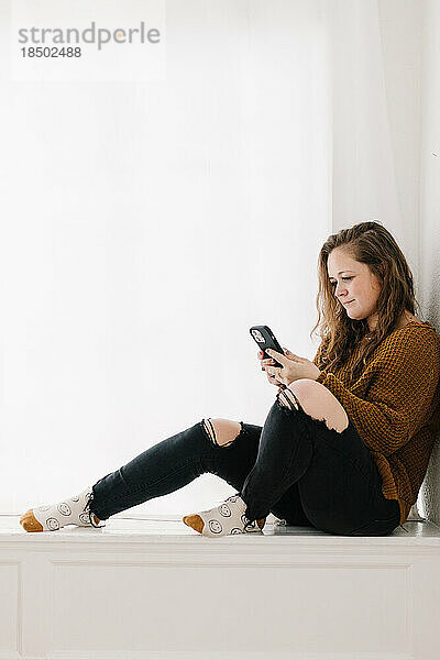 Zufriedene Frau sitzt in weißer Leseecke und schaut auf ihr Smartphone