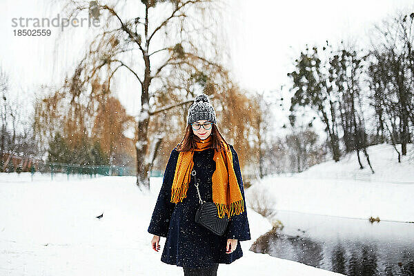 Frau in einem gelben Schal und einem blauen Mantel geht allein in einem verschneiten Park spazieren