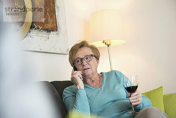 Alte Frau entspannt sich auf dem Sofa bei einem Glas Rotwein und telefoniert