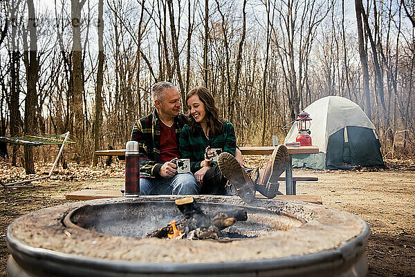 Lächelndes Paar entspannt sich im Herbst am Lagerfeuer und trinkt Kaffee
