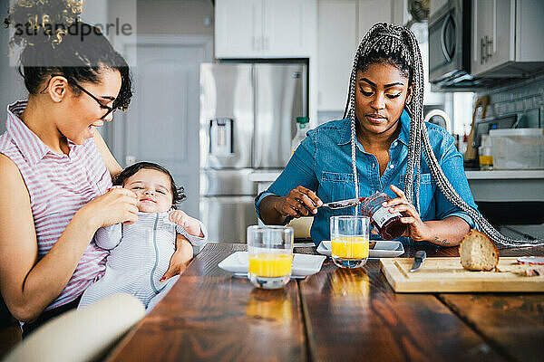 Glückliche Mutter spielt mit Baby  während Freundin am Tisch das Frühstück zubereitet