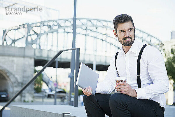 Geschäftsmann sitzt auf der Treppe und hält ein digitales Tablet mit Kaffee in der Hand  München  Bayern  Deutschland