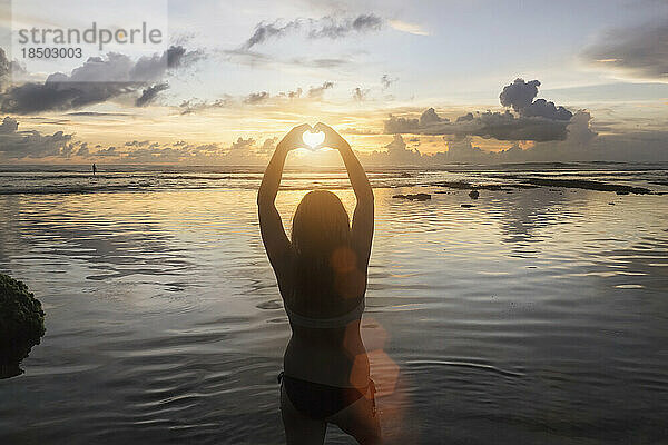 Silhouette einer Frau  die bei Sonnenuntergang über dem Meer eine Herzform macht  Uluwatu  Bali  Indonesien
